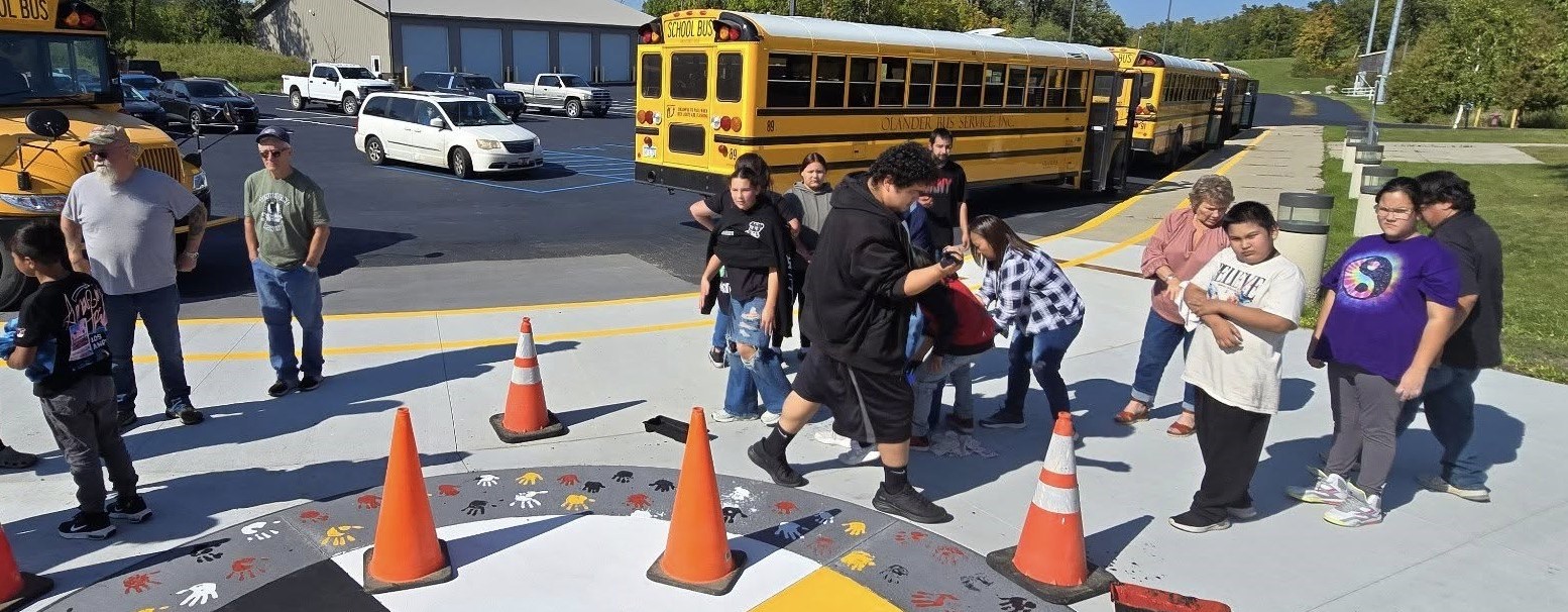A gathering of students and others outside the school with parked buses in the background.